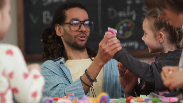 Chest up handheld shot of young biracial male teacher having art class with playful Caucasian children, sitting at table in classroom in front of blackboard and using modeling clay