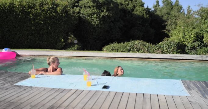 Diverse gals in bikinis seeing cam shadow, woman jumping into pool, surfacing, joining them, drinks