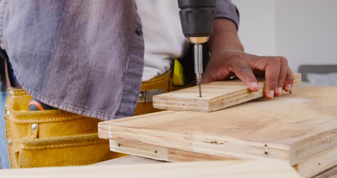 African American man assembling planks, holding screw, using cordless drill on bench with tool belt