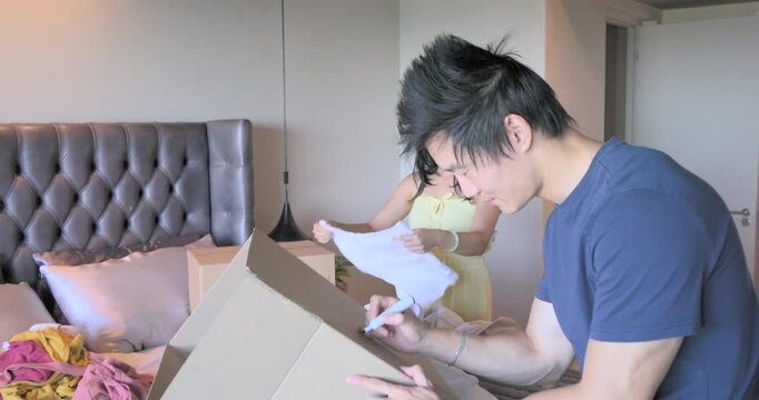Diverse couple spotting empty box, sorting and folding clothes, labeling moving boxes on bed