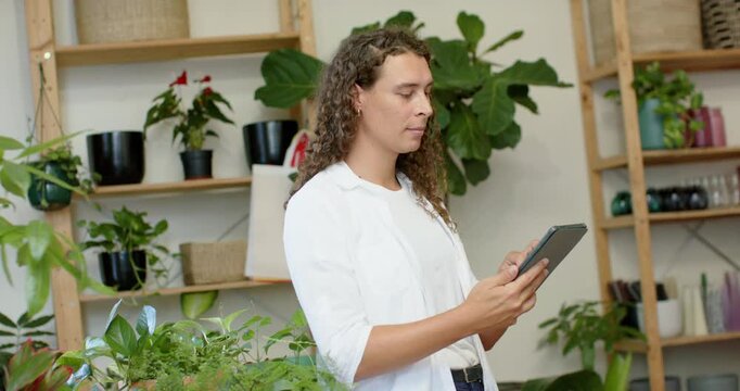 Nonbinary adult in white shirt checking tablet tapping scrolling updating stock in plant shop