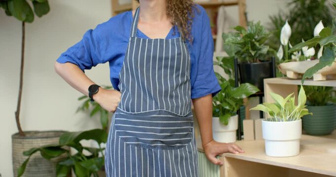 Closeup showing apron, non-binary worker adjusting strap, leaning on shelf preparing plants in shop