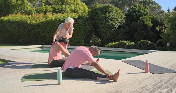 Senior African-American teacher guiding two women into forward fold on poolside mats for posture