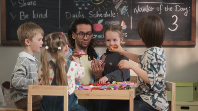 Medium slowmo shot of young biracial male teacher sitting at table with Caucasian children in front of chalkboard in kindergarten classroom and creating figures with colorful modeling clay together