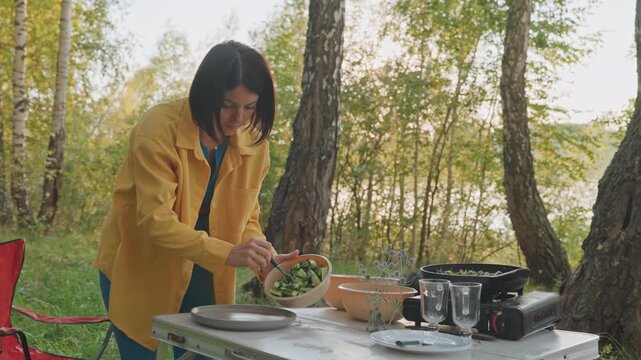 Solo camping cooking station by lake, woman in yellow jacket chops and plates fresh salad over portable stove, uses utensils and bowls on folding table, red camping chair visible, sunlight filtering