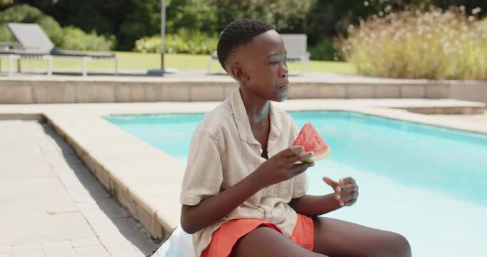African male youth sitting at backyard pool in orange shorts eating watermelon to cool in heat