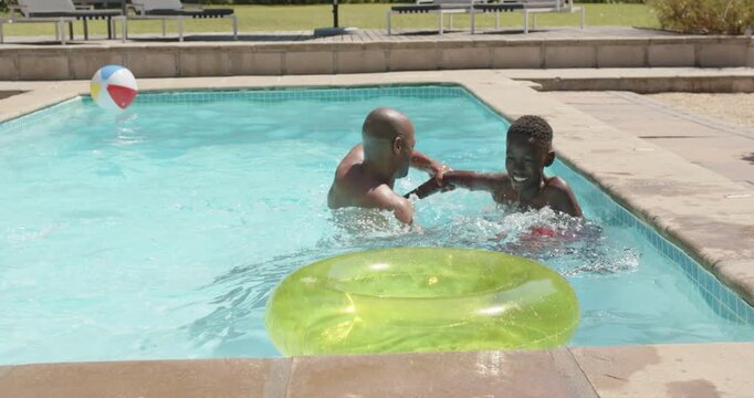 African father and youth son reaching playing tumbling in backyard pool with green ring beach ball