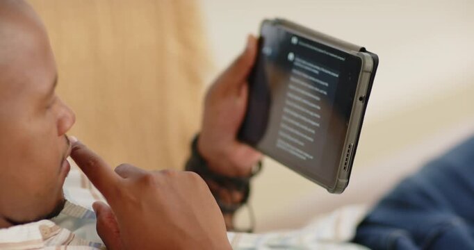 Adult man reclining on sofa holding tablet, tapping list, pausing, thinking, selecting items
