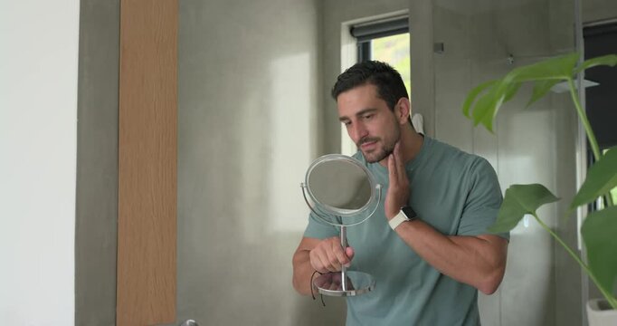 Reaching for mirror at vanity, adult man lifting and angling, checking cheeks and neck for stubble