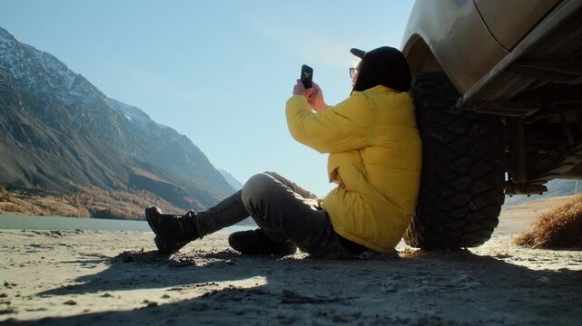 Male traveler in a yellow jacket taking a smartphone photo while sitting on the ground and leaning against an off road SUV near a mountain lake. Scenic outdoor setting with alpine slopes, open