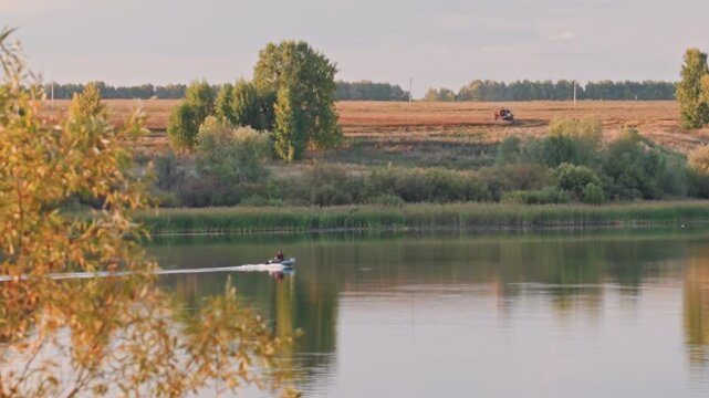 Calm river golden hour boat gliding across glassy water, rural fields and trees lining opposite bank, warm sunlight creating soft golden reflections, distant harvest activity on horizon, tranquil