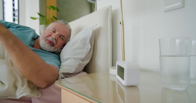 Senior couple waking in bed as digital clock ringing man reaching silencing alarm and holding her