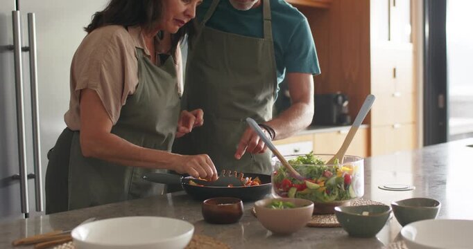 Turning vegetables in pan, couple offering bite and tasting checking flavor at island with aprons