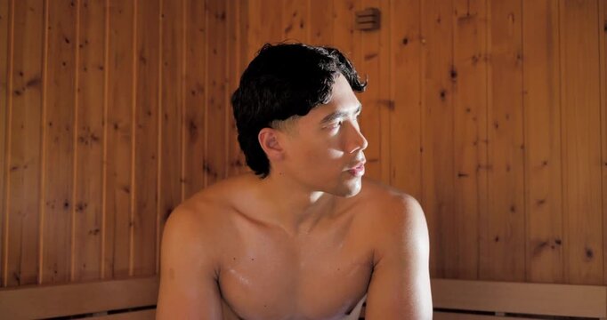 Asian man sitting on bench with towel in sauna, seeing window, turning, wiping face, breathing slow