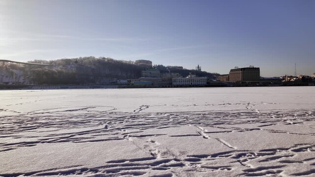 Static wide landscape of frozen Dnieper river covered in snow in Kyiv. View of Podil embankment with city buildings and smoking chimneys under clear blue sky.
