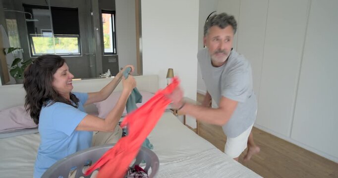 Couple responding to laundry basket, folding and passing shirts on bed in bedroom to tidy clothes