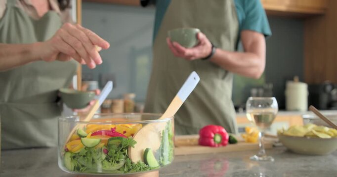 Couple chopping pepper, sprinkling, stirring glass bowl on kitchen island in aprons preparing salad