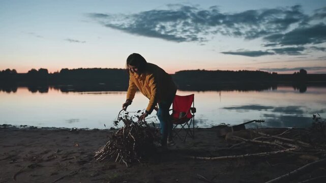 Twilight lakeside camping woman arranging kindling and tossing branches onto pile by calm water, red folding chair behind, motion and purpose as campsite is prepared at dusk, reflective sky and quiet