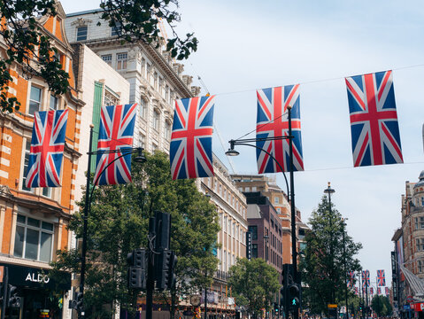London, United Kingdom - 20 June 2025: View of Union Jack flags fluttering above a bustling London street, casting shadows on the vibrant facades and lush green trees below.