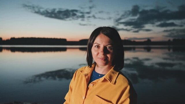 Smiling woman by lake at dusk camping, closeup portrait in yellow jacket with soft twilight horizon, warm light on face and calm water backdrop, friendly camper energy and relaxed evening atmosphere