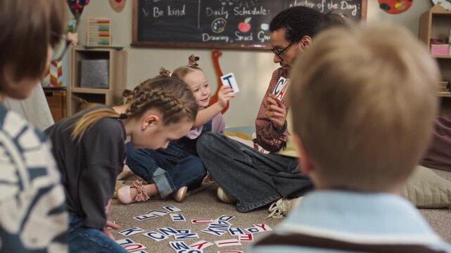 Handheld slowmo shot of motivated Caucasian girl child sitting on floor in cozy kindergarten classroom among her peers near young biracial male teacher and learning alphabet using flashcards