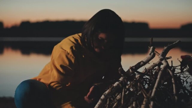 White woman preparing camping tinder with hands close to lighter at lakeshore dusk, arranging small sticks and kindling, focused closeup, reflective mood, calm twilight, firecraft technique, outdoors
