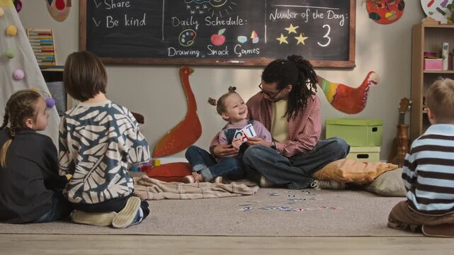 Ground level slowmo shot of caring young biracial male kindergarten teacher sitting on floor and interacting with joyful Caucasian girl holding flashcards in cozy classroom among other children
