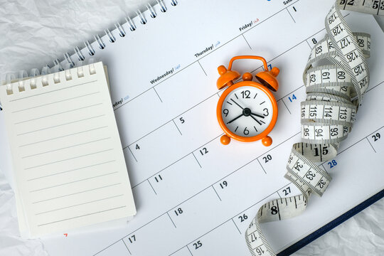 Blank notepad with an orange alarm clock and measuring tape on a monthly calendar background. Suitable for meal planning notes and fitness tracking designs.