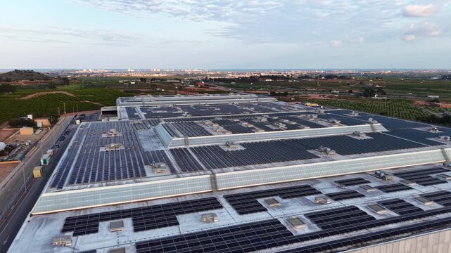 Large industrial warehouse rooftop covered with photovoltaic panels surrounded by agricultural fields and a distant cityscape