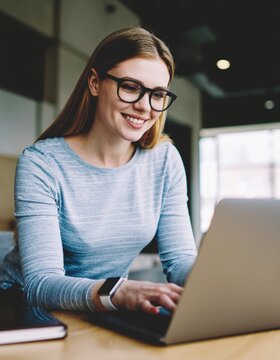 Young smiling woman wearing glasses using laptop at a modern cafe, enjoying remote work and online learning experience