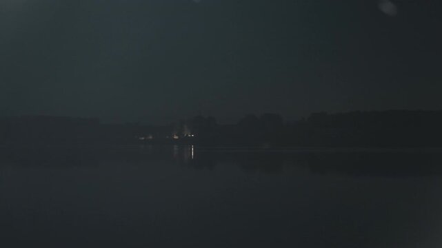 Night lake camping scene with distant lights, soft snow flurries drifting across calm water, lone cabin glow on opposite shore, silhouetted trees, subtle reflections, camper holding lantern, angler