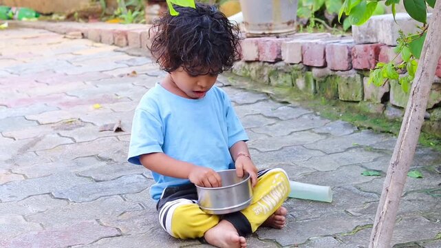 A cute little Indian toddler girl sitting barefoot on an outdoor paved courtyard, independently eating from a small stainless steel bowl placed between her legs, capturing an authentic moment 