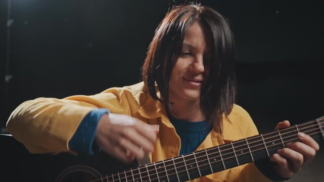Smiling white woman playing guitar, camping musician in yellow jacket, playful finger gesture and tongue out expression, candid studio lighting, welcoming singalong vibe with unplugged folk energy