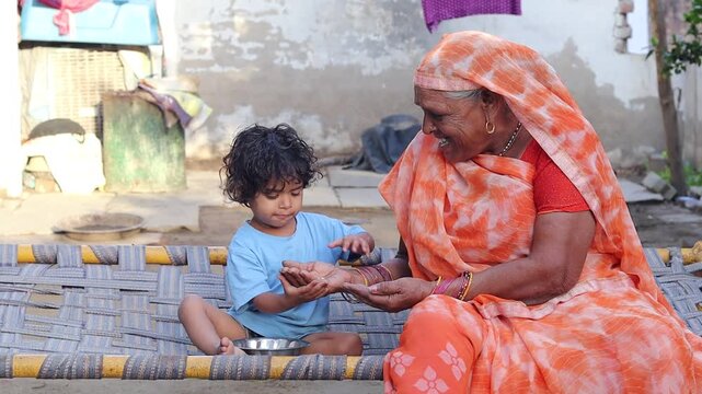 An affectionate elderly Indian grandmother sitting on a traditional woven cot (charpai) outdoors, lovingly watching her toddler grandchild eat from a small stainless steel bowl, highlighting rural fam