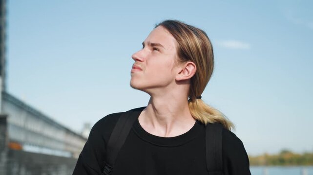 Young individual looks sideways. Youthful person glances to side. Young man with backpack gazes sideways attentively. Youthful individual carrying backpack observes objects passing by on side