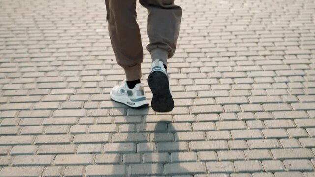 Caucasian man walking on cobblestone pavement wearing white sneakers, casual brown pants, long shadow from low sunlight, brisk stride and relaxed urban atmosphere, closeup on footwear and textured