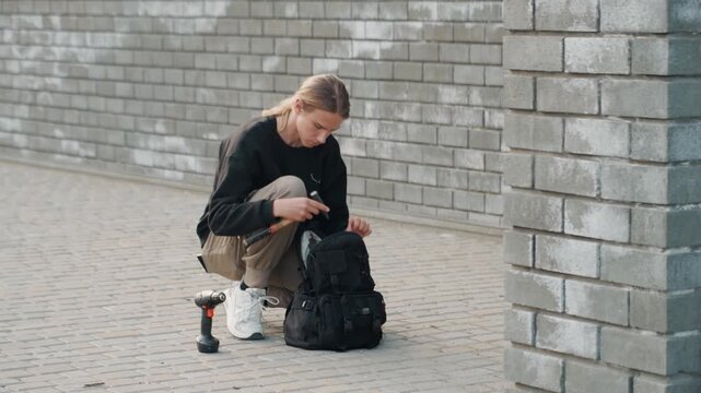 Young person kneeling by backpack inspecting gimbal and camera gear against brick wall, quiet urban setting, focused preparation for street shoot, subtle light and textured pavement
