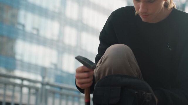 Young white man checking phone and tools before departure. Final gear check at waterfront, secures equipment in backpack, thoughtful commute across city promenade with skyline backdrop and quiet