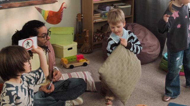 Handheld shot of joyful Caucasian children playing in cozy kindergarten classroom while learning road signs on flashcards during educational activity with young biracial male teacher sitting on floor