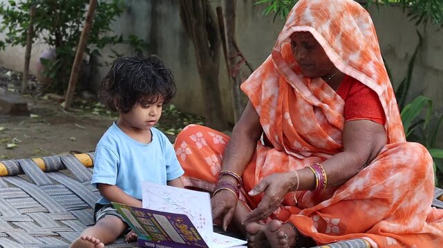An affectionate elderly Indian grandmother and her toddler grandchild sitting together on a traditional woven cot (charpai) outdoors. The child is looking at an open book, depicting early childhood ed