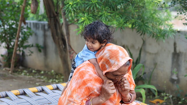 Candid shot of a loving Indian grandmother carrying a toddler on her back, sitting on a charpai in a rural courtyard.