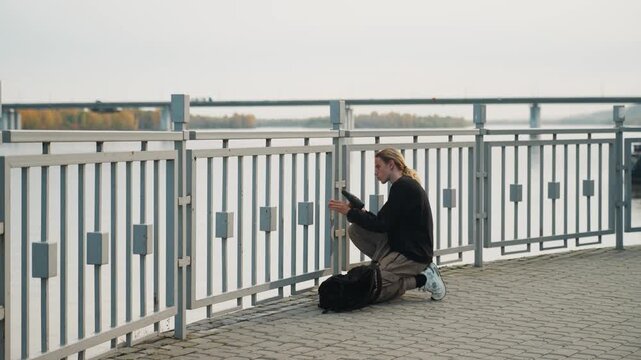 Photographer reviewing photos. Kneeling photographer calibrates device near river. Focused young photographer crouches by water reviewing images on their device in serene autumn surroundings