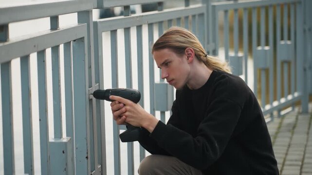 Young white man installing sensor on bridge railing, kneeling while securing mount with drill, concentrated expression, braided hair, casual jacket and backpack, waterfront skyline, urban maintenance