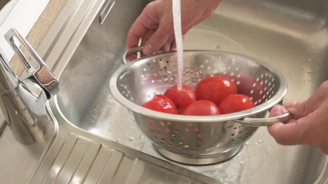 Washing fresh tomatoes in a large colander under running water in kitchen
