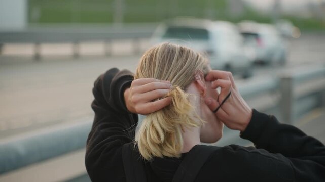 Blonde white woman tying hair overpass, commuter preparing for evening shift, hands gather strands and fingers secure elastic, focused styling gesture, blurred cars, candid back view