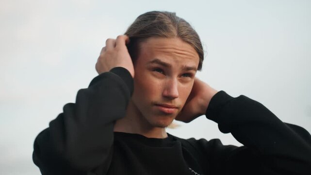 Young man adjusting hair on overpass, backpack and black sweater, soft dusk sky, contemplative gaze and gentle hand movements smoothing long hair, candid closeup portrait with natural light