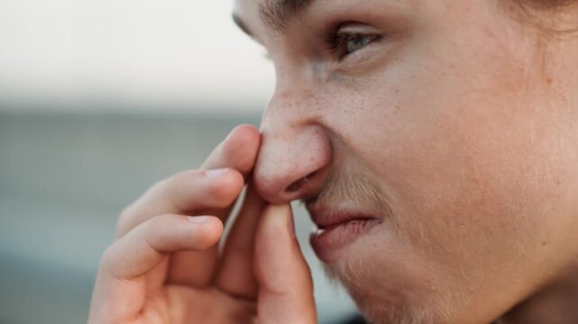 White teen pinching nose outdoors, reacting to strong odor on waterfront, freckled skin and light stubble, closeup profile shot, hand pressing nostrils, tense expression, candid natural lighting,