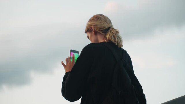 Caucasian woman streaming under cloudy sky, low angle view with dramatic clouds, hands framing phone while recording, tension and focus, broadcast and realtime engagement, moody urban atmosphere