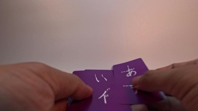 A close-up shot captures a person&rsquo;s hands flipping through purple educational flashcards to reveal Japanese Hiragana and Katakana characters, starting with the vowels A, I, U, E, and O