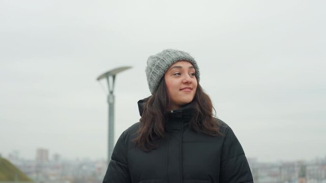 Woman in gray beanie smiling with playful dancelike gestures near lamppost, city skyline backdrop, curious upward gaze, candid urban portrait, chilly coat and knit hat, city explorer energy